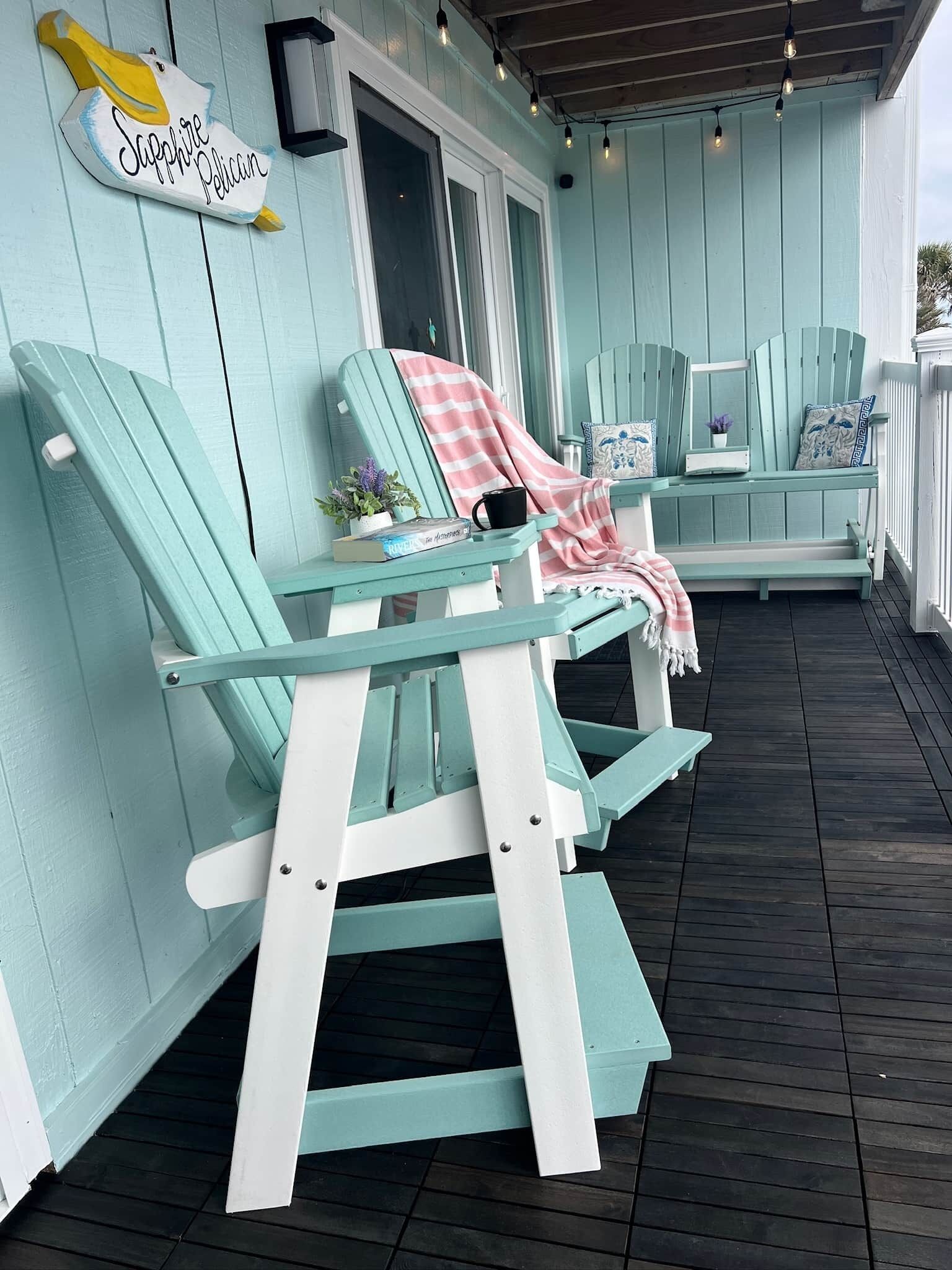 A covered outdoor seating area with turquoise Adirondack chairs, a sign reading "Sapphire Pelican," and a string of lights above.