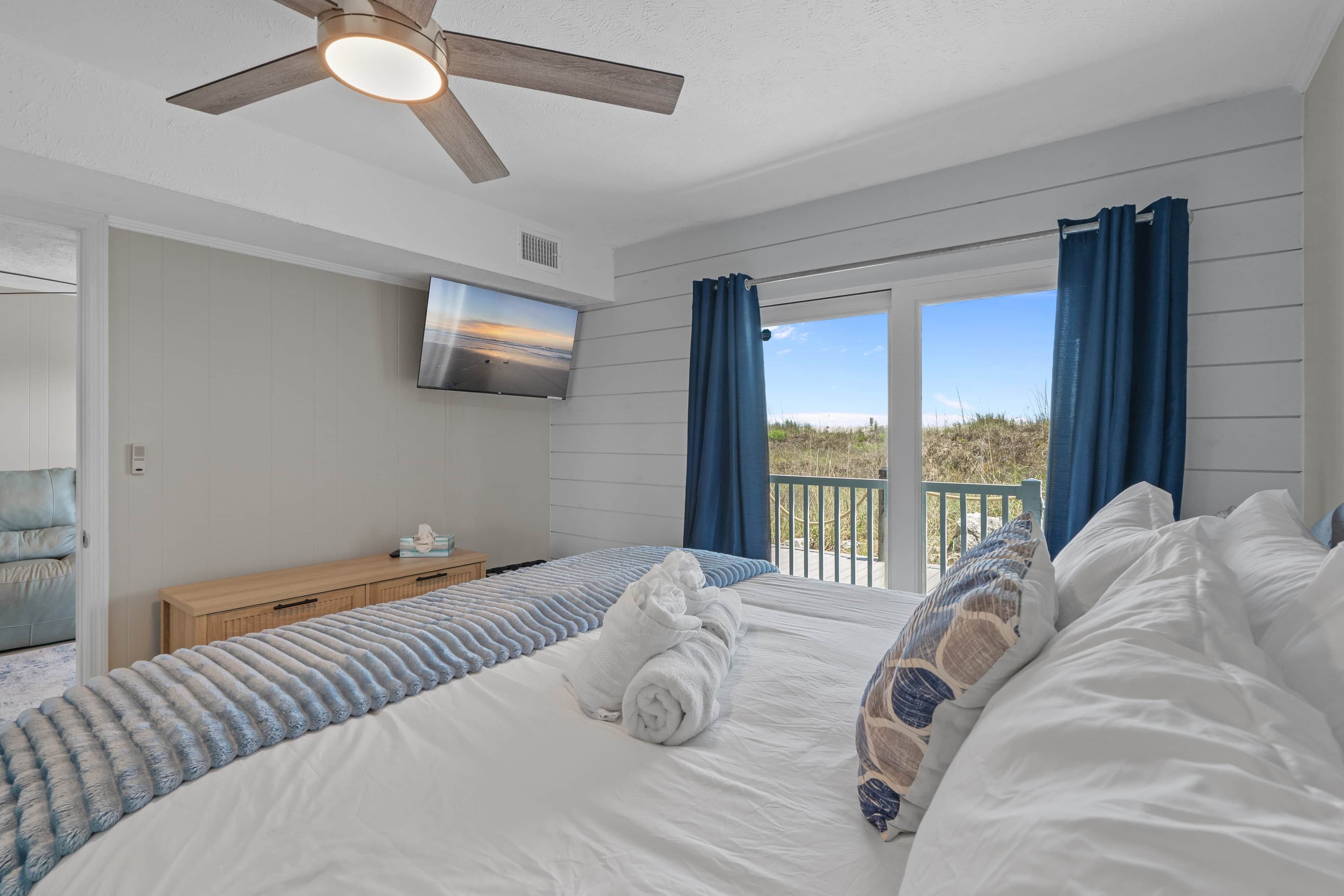 A bedroom with a bed, a ceiling fan, a TV, and a view of the outdoors.