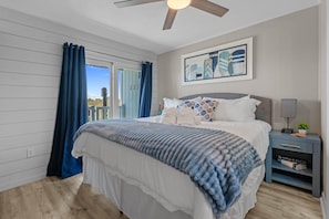 A bedroom with a bed, a nightstand, a ceiling fan, a window with curtains, and a wall-mounted painting.