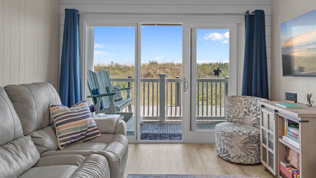 A living room with a sofa, a patterned armchair, a bookshelf, and a sliding glass door leading to a deck.