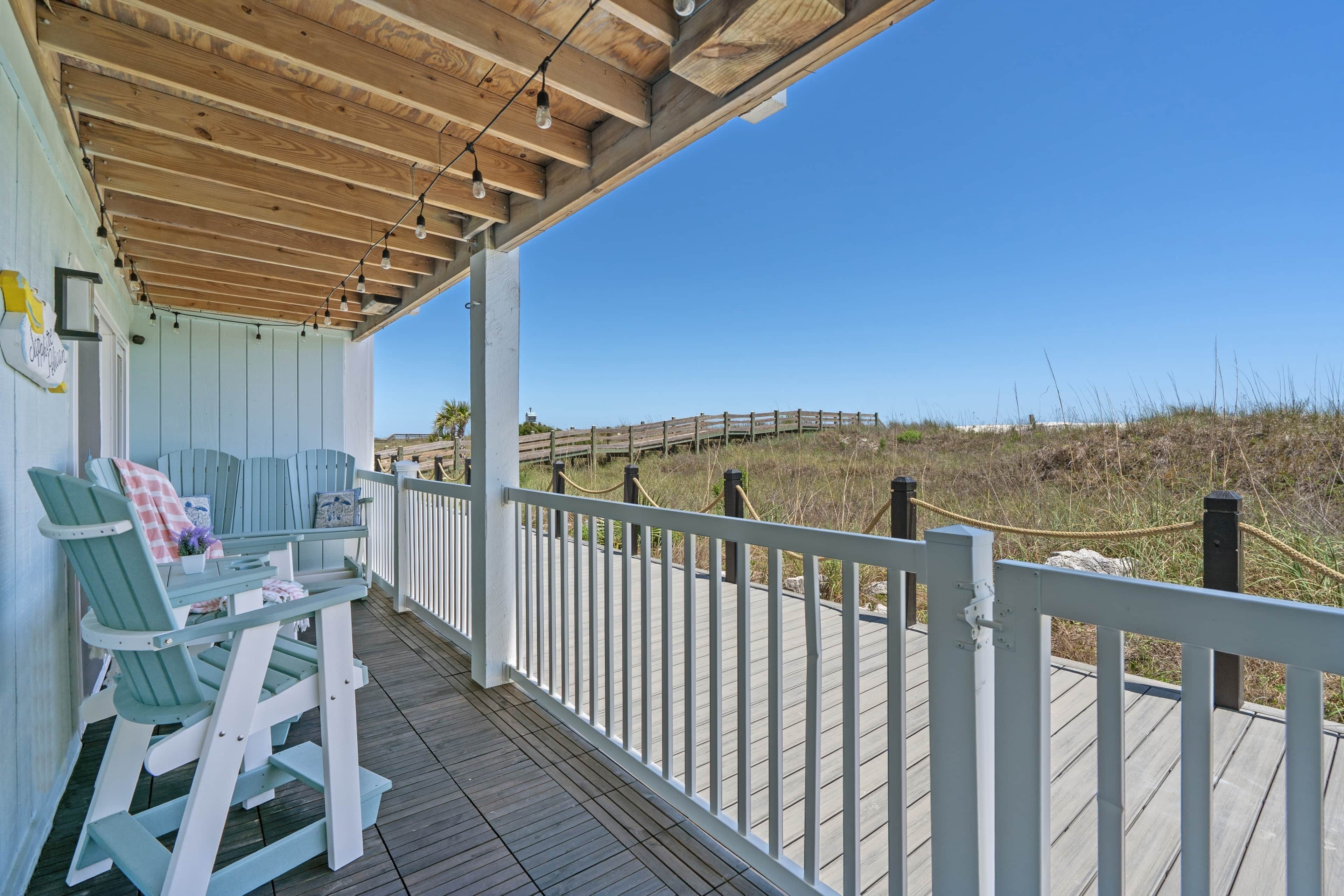 A wooden deck with a white railing, green chairs, and a view of the beach.