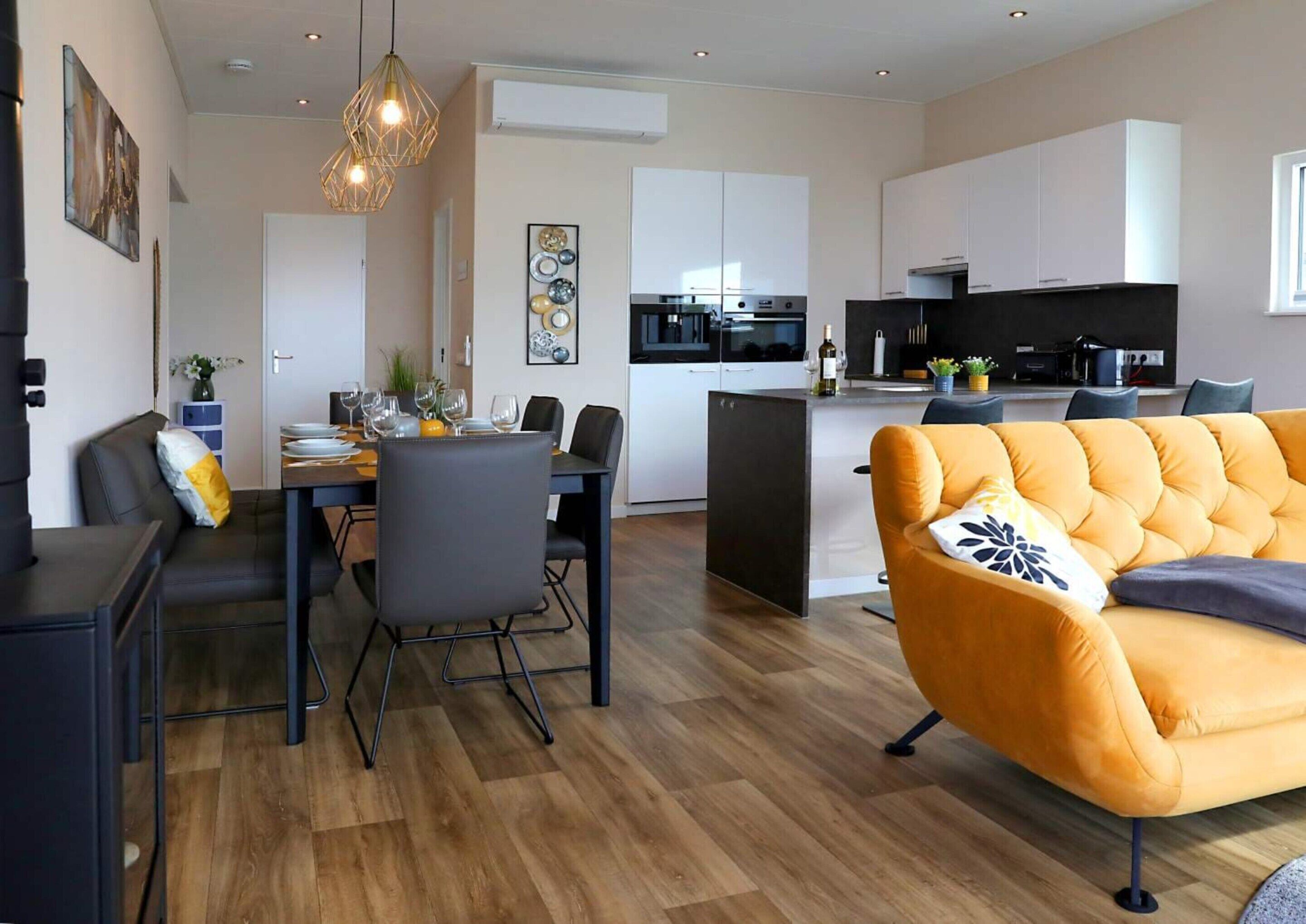 A modern living room with a yellow sofa, dining area with a table and chairs, and a kitchen with white cabinets and a black countertop.