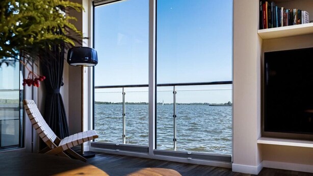 A modern room with a large window overlooking a body of water, a lounge chair, a small table, and a bookshelf. - Coastal Comfort With Terrace (Ribnitz-Damgarten)