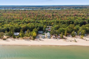 An aerial view of a coastal area with a beach, a cluster of houses surrounded by dense forest, and a distant shoreline.
