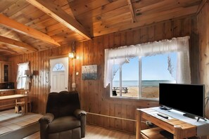 A wooden cabin interior with a TV, a wooden desk, a chair, a dining table, and a view of the beach.