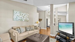 A living room with a beige sofa, a brown ottoman, a flat-screen TV, and a painting on the wall.