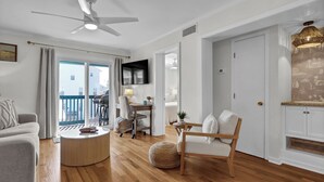 A modern living room with a ceiling fan, a flat-screen TV, a wooden coffee table, a sofa, a chair, and a dining area with a table and chairs. - Carolina Beach Retreat | Pool, Ocean View, Beach Access & Stylish Decor (Carolina Beach)