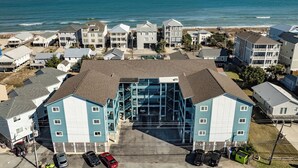 An aerial view of a coastal hotel with multiple buildings, a parking lot, and a beach in the background. - Carolina Beach Retreat | Pool, Ocean View, Beach Access & Stylish Decor (Carolina Beach)