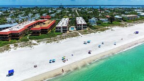 Luftaufnahme eines Strandresorts mit mehreren Gebäuden, einem Sandstrand und klarem türkisblauem Wasser.