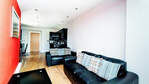 A modern living room with a black leather sofa, a dining area with a wooden table, and a kitchen in the background. - 2 Bedroom Apartrment (Newcastle-upon-Tyne)