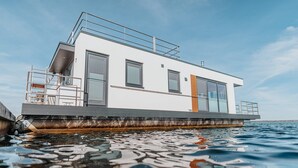 A modern floating house with a flat roof, large windows, and a deck area.