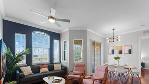 Una sala de estar moderna con una pared de acento azul oscuro, un ventilador de techo, una zona de comedor con mesa y sillas, y un ventanal con persianas.