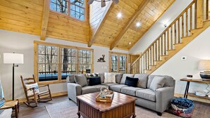 A spacious living room with a wooden ceiling, large windows, a gray sofa, a wooden coffee table, and a staircase.