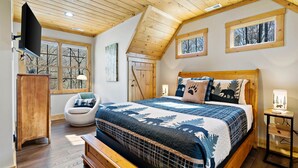 A bedroom with a wooden ceiling, a large bed with patterned bedding, a chair, and a desk with a lamp.