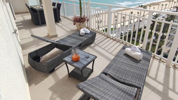 Ein Balkon mit Korbmöbeln, einem Kaktus und Blick auf Strand und Meer.