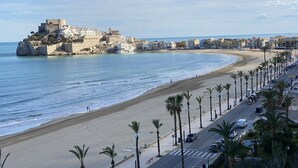 Eine Küstenstadtlandschaft mit einer markanten historischen Festung auf einem Hügel, einem breiten Sandstrand und einer von Bäumen gesäumten Promenade.