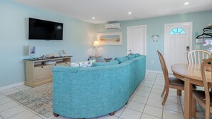 Una sala de estar con un sofá color turquesa, un televisor de pantalla plana, una mesa de comedor con sillas y una alfombra pequeña.