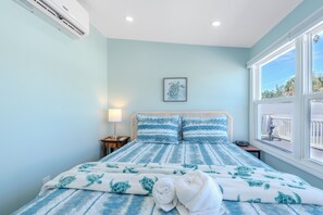 Un dormitorio con cama, mesita de noche, lámpara, un cuadro enmarcado en la pared y ventana con vistas al exterior.