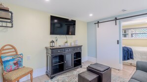A modern living room with a flat-screen TV mounted on the wall, a wooden chair with a colorful cushion, a dark wooden cabinet with drawers, and a gray sofa.