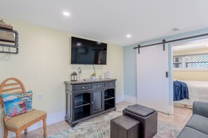 A modern living room with a flat-screen TV mounted on the wall, a wooden chair with a colorful cushion, a dark wooden cabinet with drawers, and a gray sofa.