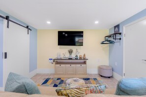 Een woonkamer met een flatscreen televisie aan de muur, een klein kastje met decoratieve objecten, een kamerkleed en een beklede bank.