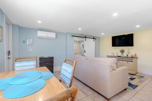 Ein modernes Wohnzimmer mit einer Couch, einem Esstisch mit Stühlen, einem an der Wand montierten Fernseher und einer Klimaanlage.