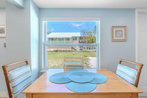 Un comedor con una mesa y sillas de madera, vista de una casa a través de un amplio ventanal y un cuadro en la pared.