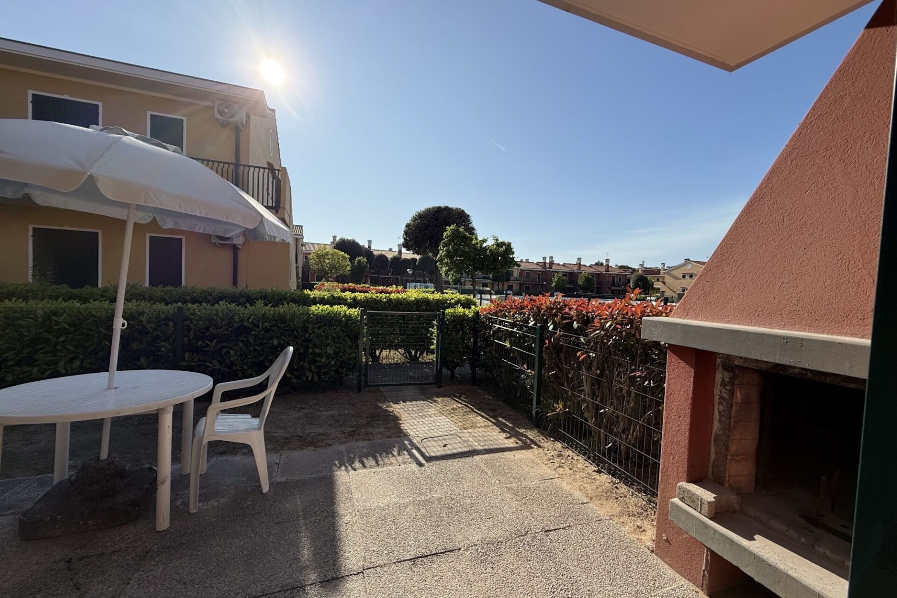 Um pátio com mesa e cadeiras, um guarda-sol branco, um forno de tijolos e vista para os edifícios e vegetação ao redor.