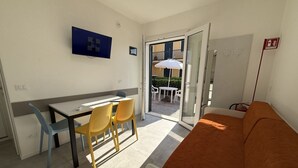 A room with a dining table, chairs, a sofa, a TV, and a sliding glass door leading to an outdoor area with a table and chairs. - Cozy Apartment With Private Garden in Bibione (San Michele al Tagliamento)