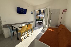 Ett rum med ett matbord, stolar, en soffa, en tv och en skjutdörr i glas som leder ut till ett utomhusområde med ett bord och stolar.