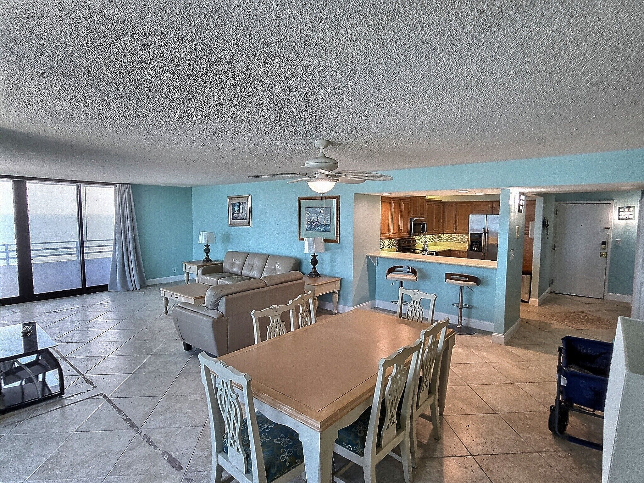 A spacious living area with a large sofa, dining table, and chairs, a kitchen with bar stools, and a balcony with a view.