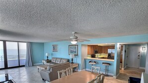 A spacious living area with a large sofa, dining table, and chairs, a kitchen with bar stools, and a balcony with a view.