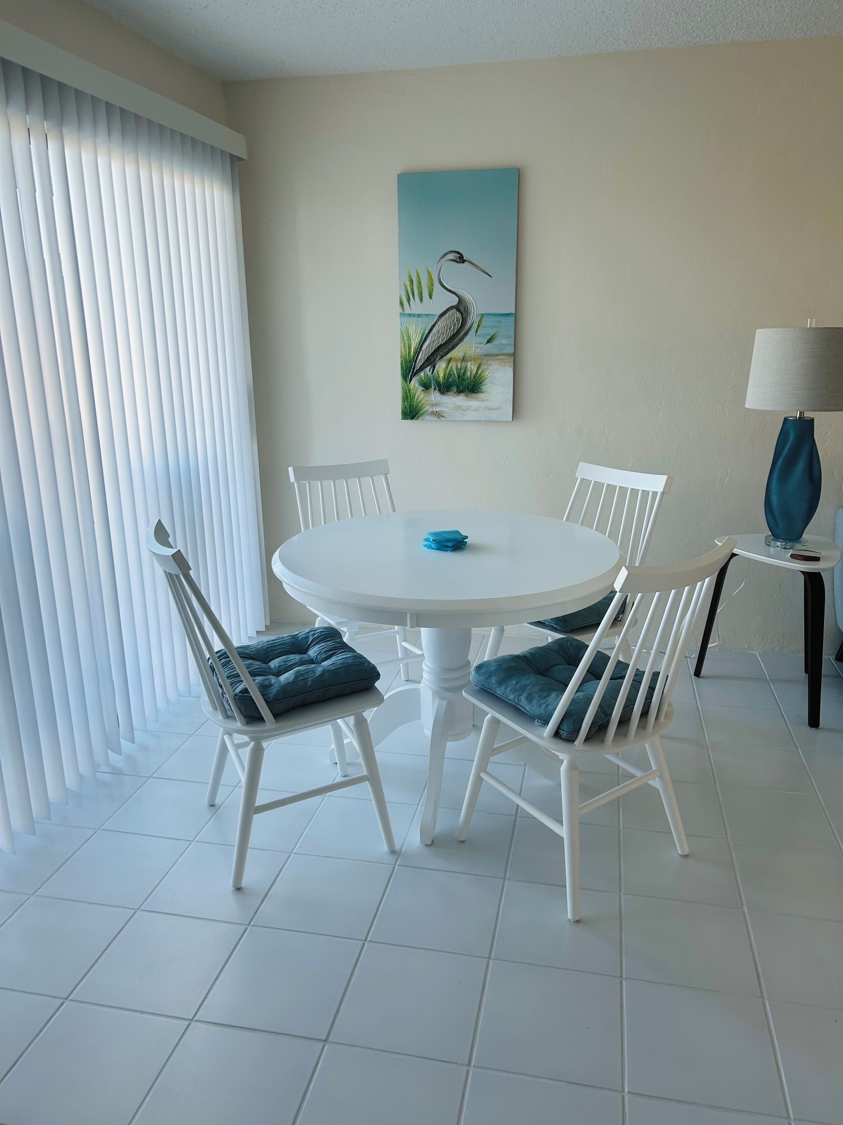 Un comedor con una mesa redonda blanca, cuatro sillas blancas con cojines azules, un cuadro de un pájaro en la pared y una lámpara sobre una mesita auxiliar al lado del comedor.