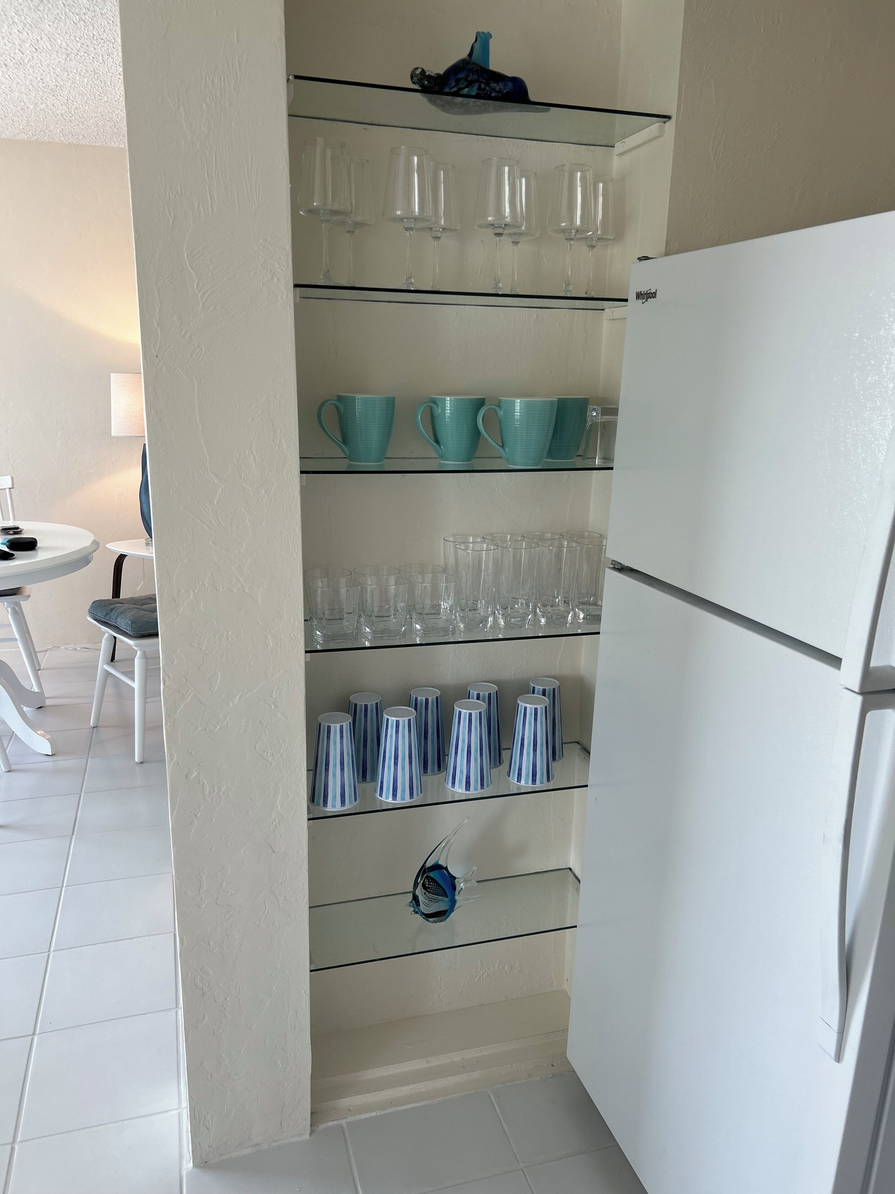 Una cocina con un refrigerador blanco, un estante de vidrio con tazas y vasos varios, y un comedor con una mesa y sillas blancas.