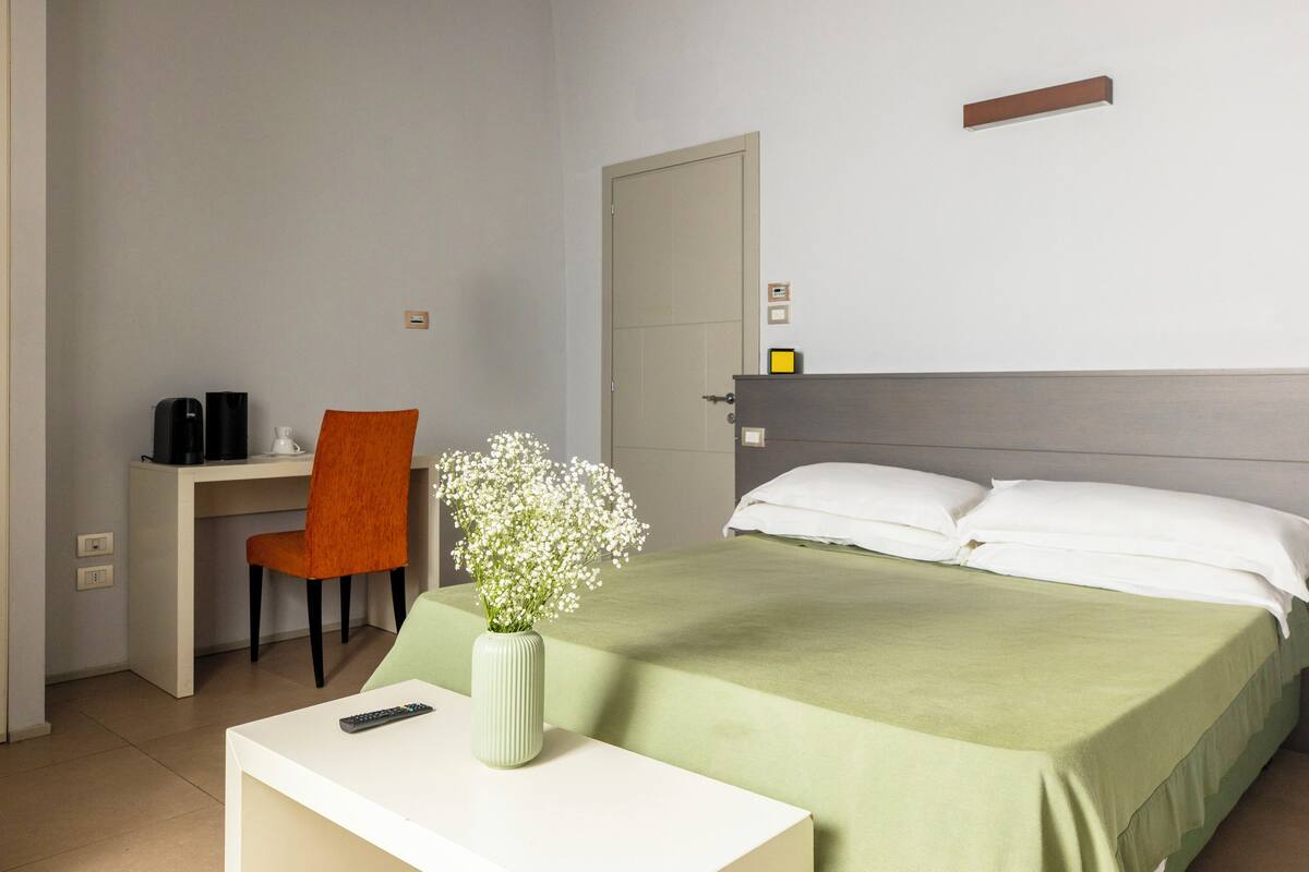 Ein Hotelzimmer mit Bett, Schreibtisch, Stuhl, einer Vase mit Blumen und einem Nachttisch.