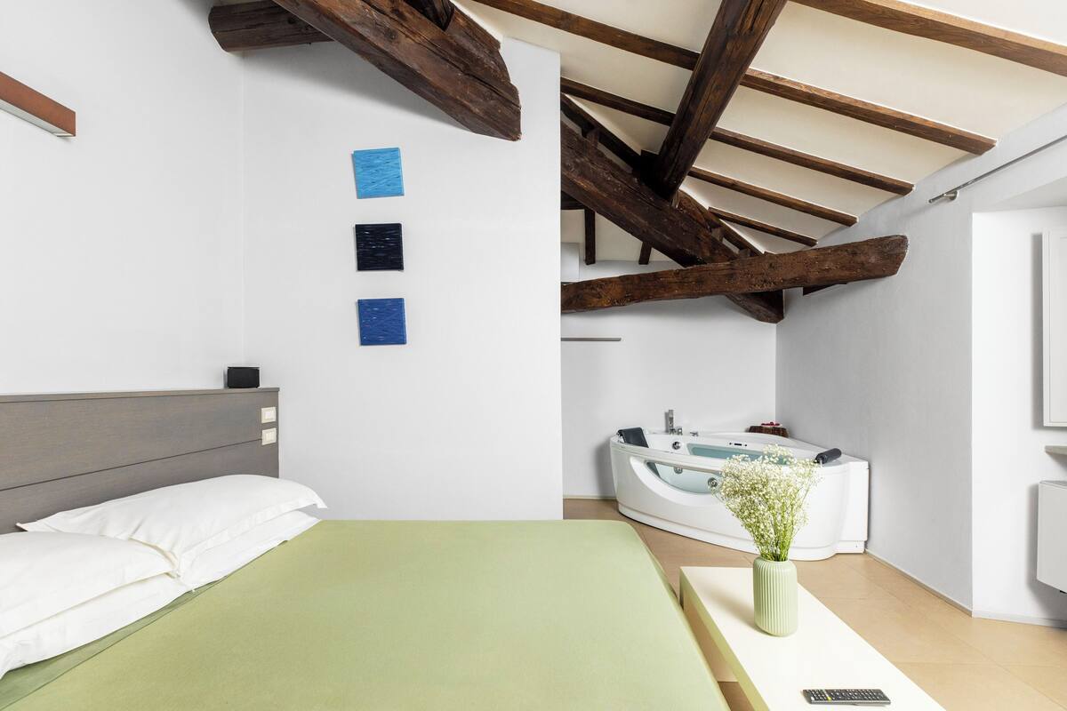 Ein Schlafzimmer mit einem Bett, einer grünen Bettdecke, einem weißen Kissen, einem Nachttisch, einer Vase mit Blumen, einer weißen Badewanne und Holzbalken an der Decke.
