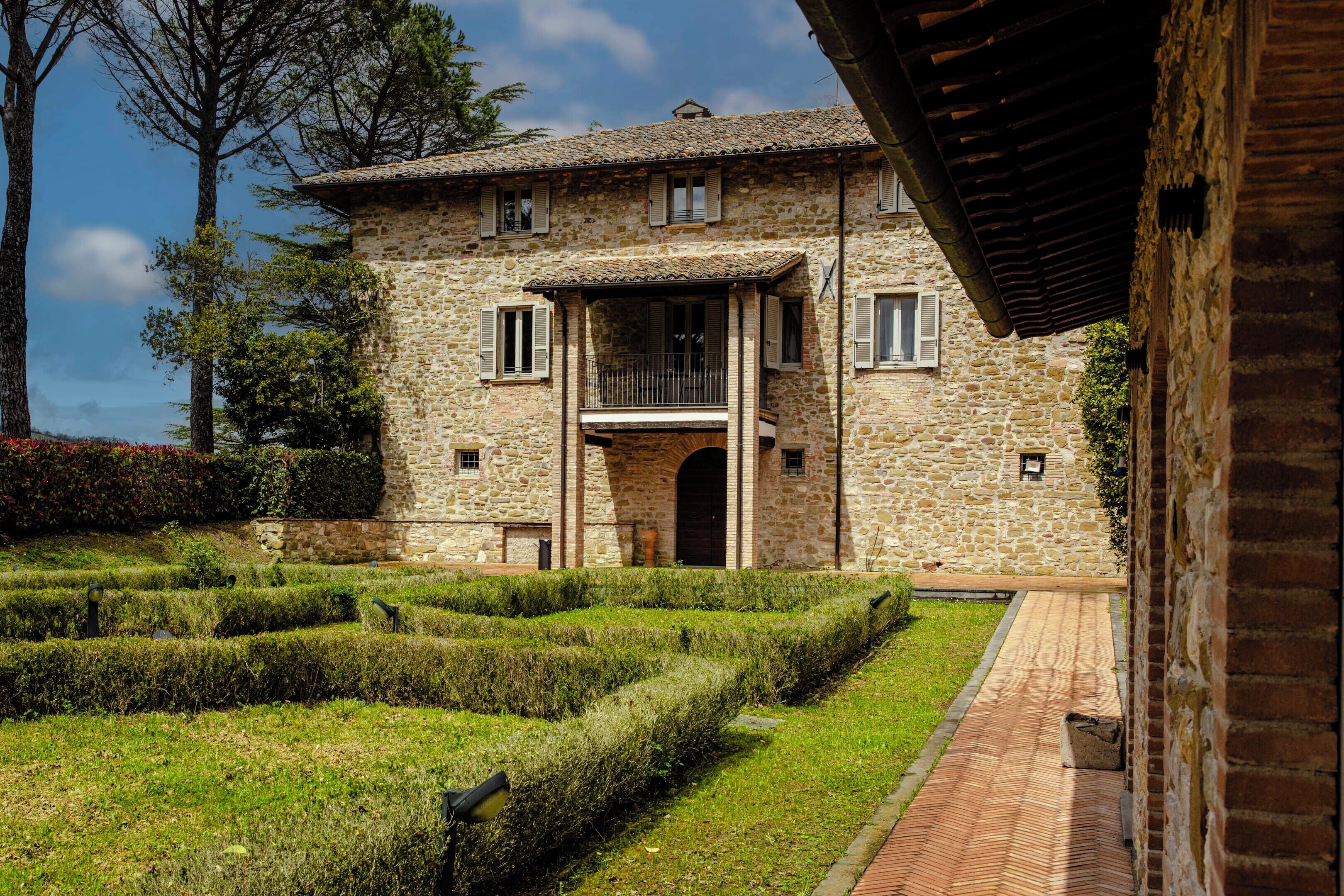 A stone building with a balcony, surrounded by a well-maintained garden with hedges and a brick pathway.