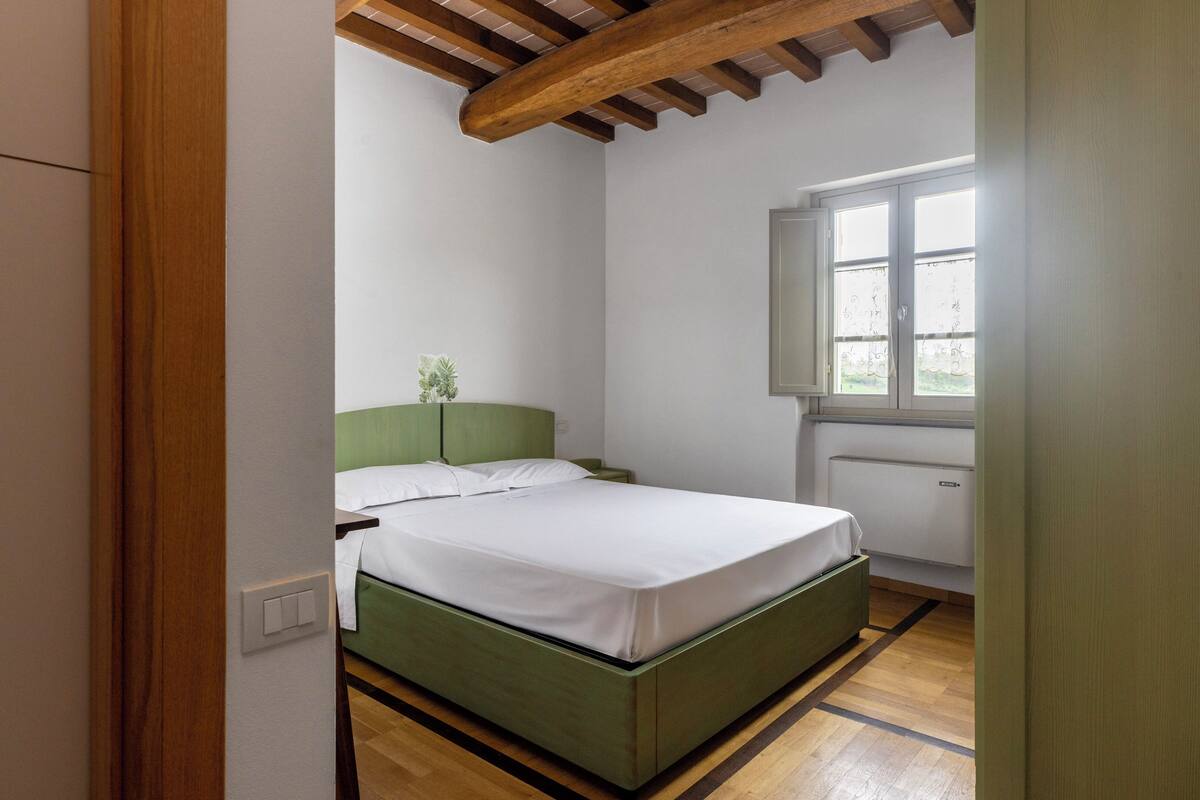 Ein Schlafzimmer mit einem grünen Bett, weißer Bettwäsche, einem Nachttisch, einem Fenster mit Vorhängen und einer Holzbalkendecke.