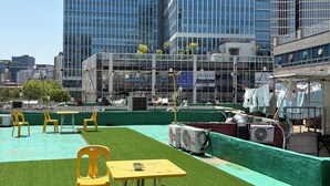 Een terras op het dak met kunstgras, gele stoelen, een tafel en airconditioningsapparaten.