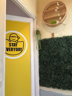 Une porte jaune avec une pancarte indiquant « RESTEZ TOUS LES JOURS », un cadre de porte blanc, un mur verdoyant couvert de feuilles et une étagère en bois sur laquelle est posée une plante.