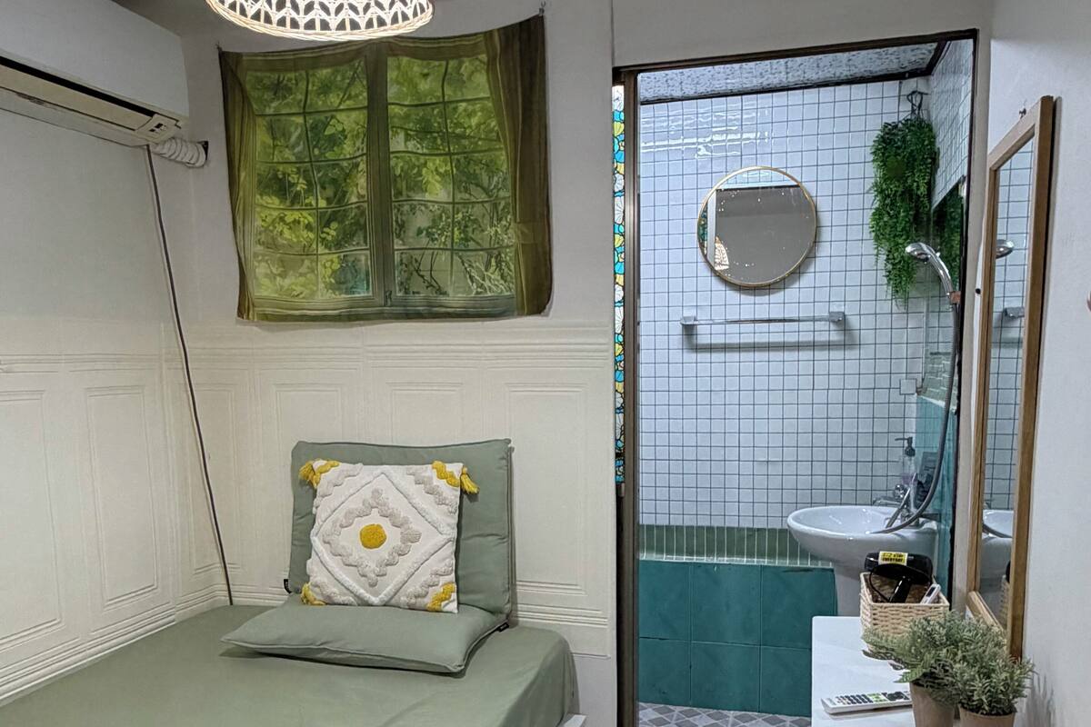 A small, modern bedroom with a bed, a hanging light fixture, a window with a view, a bathroom with a sink and mirror, and a small table with a plant.