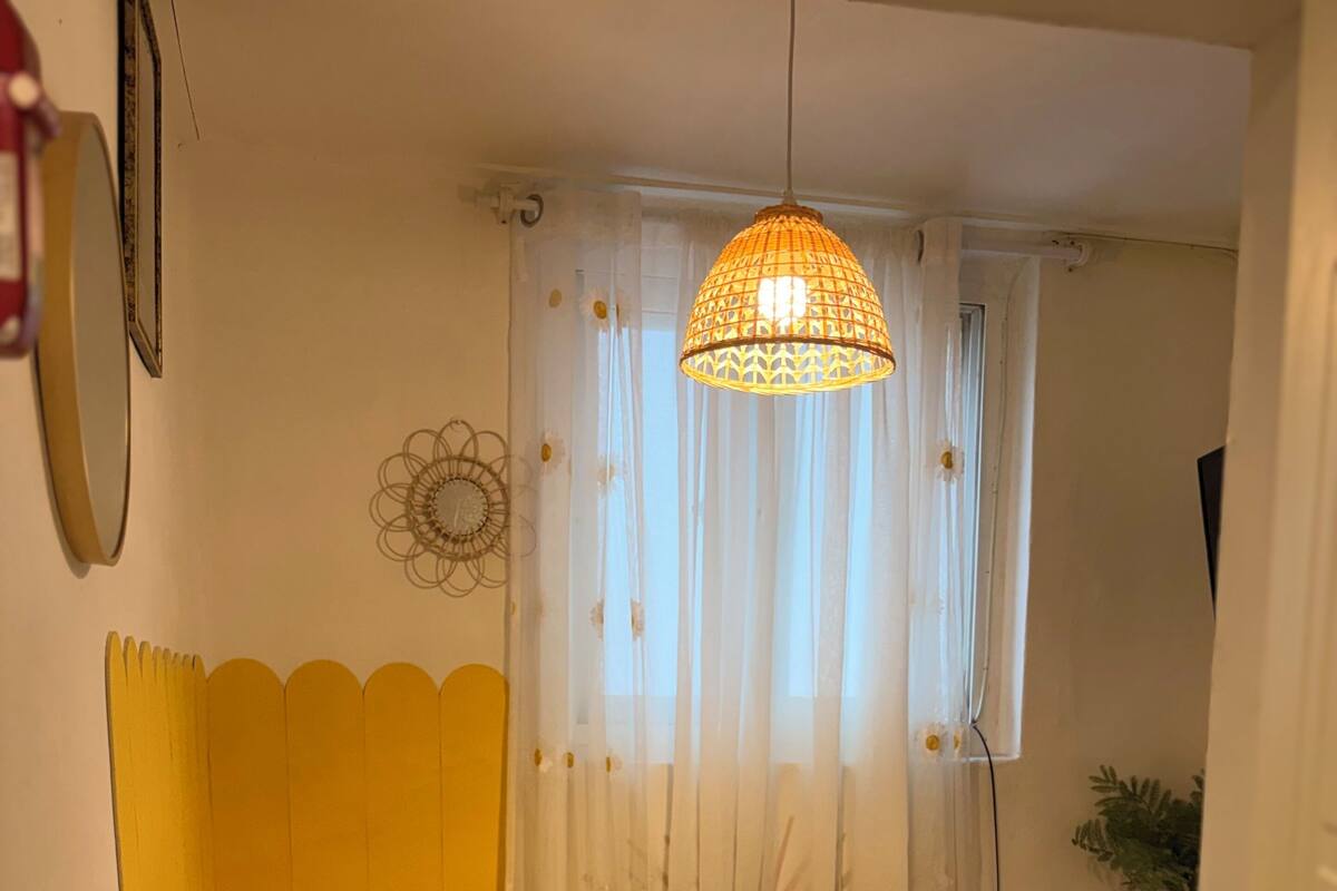 A small, cozy bedroom with a bed, a bedside table, a hanging light fixture, and a window with sheer curtains.
