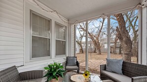 Une véranda couverte avec des meubles en osier, une table en verre ornée d’un vase de fleurs et une vue sur un jardin avec des arbres et une maison.