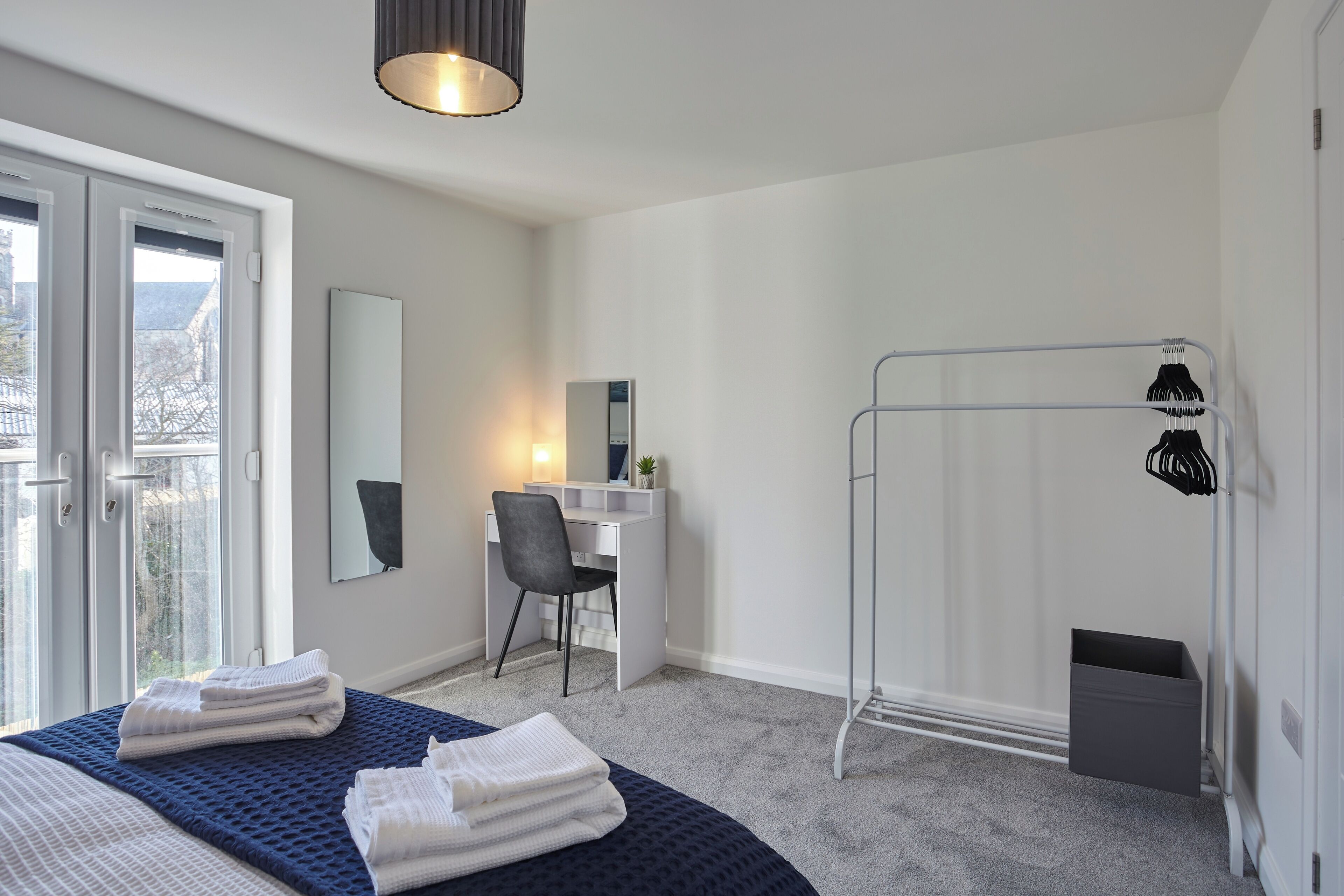 Ein modernes Schlafzimmer mit einem großen Bett, einem Schreibtisch und einem Stuhl. Ein Fenster bietet Blick auf Gebäude.