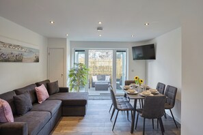 A modern living room with a sofa, dining table, and chairs. There is a TV mounted on the wall and a painting on the wall. - 2022 Modern Home In Central Hayle With Parking (Hayle)