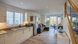 Cocina moderna con armarios blancos, refrigerador de acero inoxidable y microondas integrado. Se ve un sofá oscuro y, al fondo, un comedor con mesa y sillas.