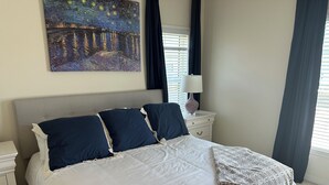 Un lit bien fait, avec une couette blanche et des oreillers bleu marine, une table de chevet avec une lampe, et un tableau représentant une scène nocturne accroché au mur.