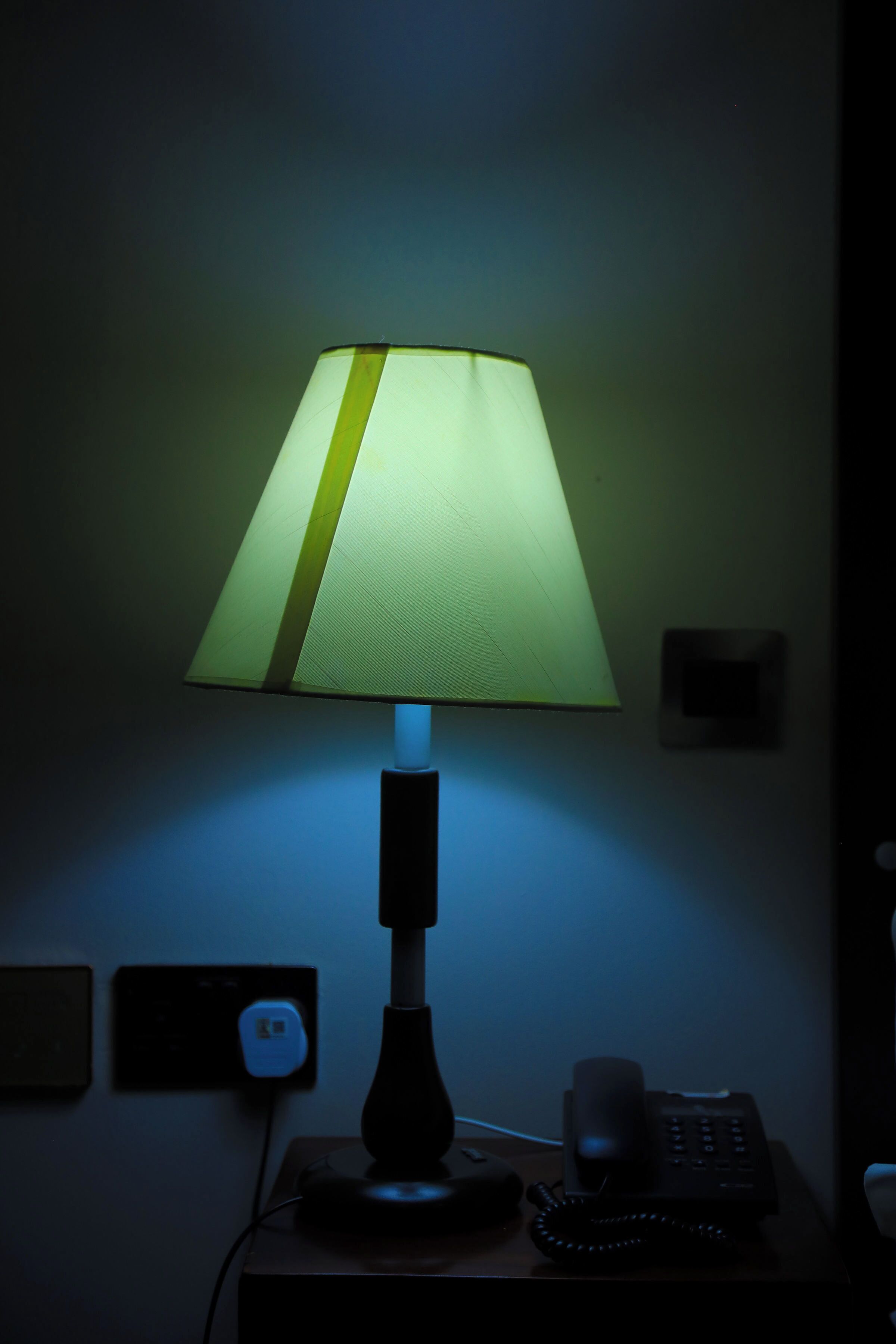 A table lamp with a green shade on a bedside table next to a black telephone.