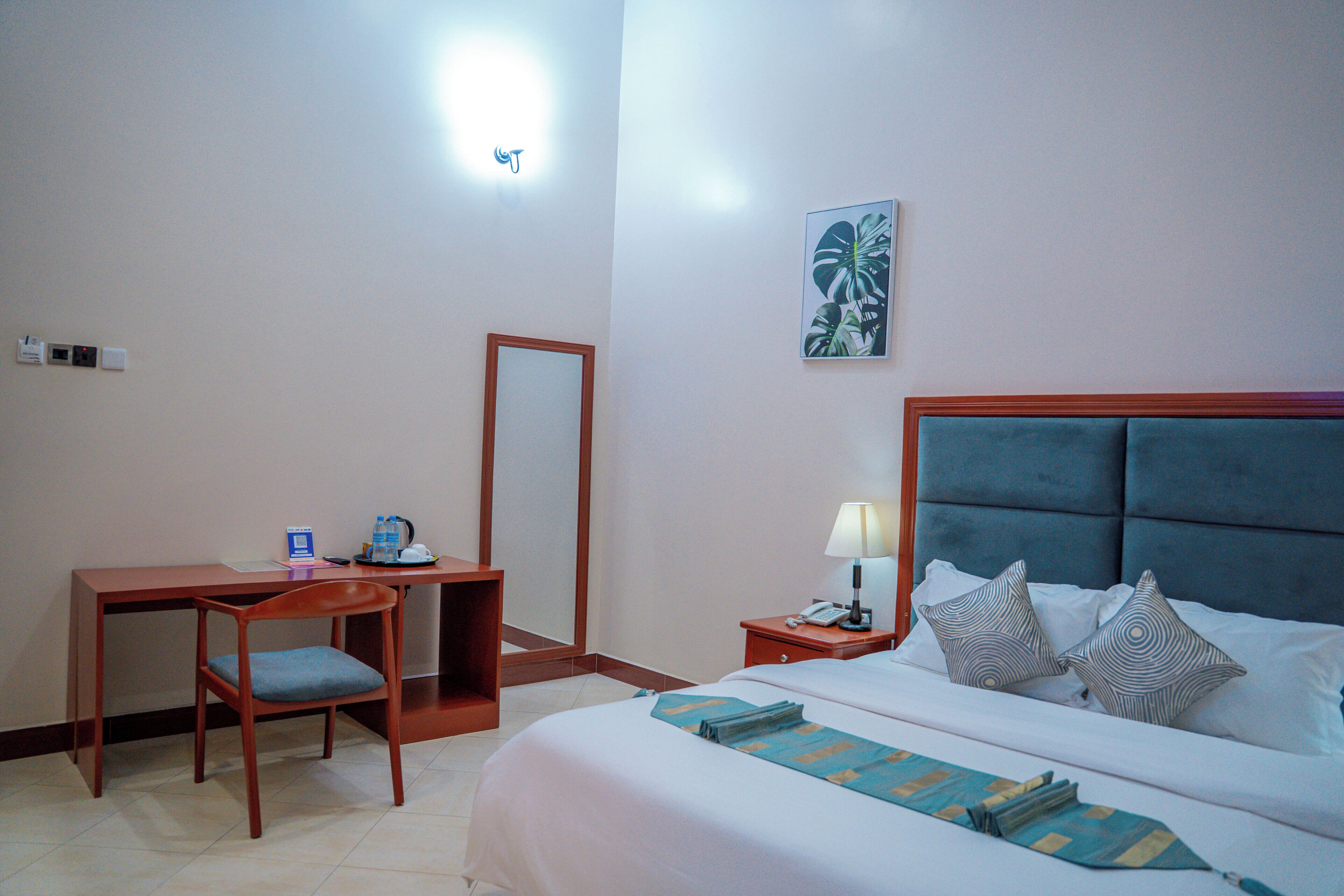 Habitación de hotel con cama, escritorio con silla, mesita de noche con lámpara y un espejo grande en la pared.
