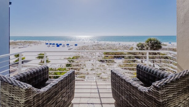 Een balkon met rieten meubilair met uitzicht op een strand met blauwe stoelen en palmbomen.
